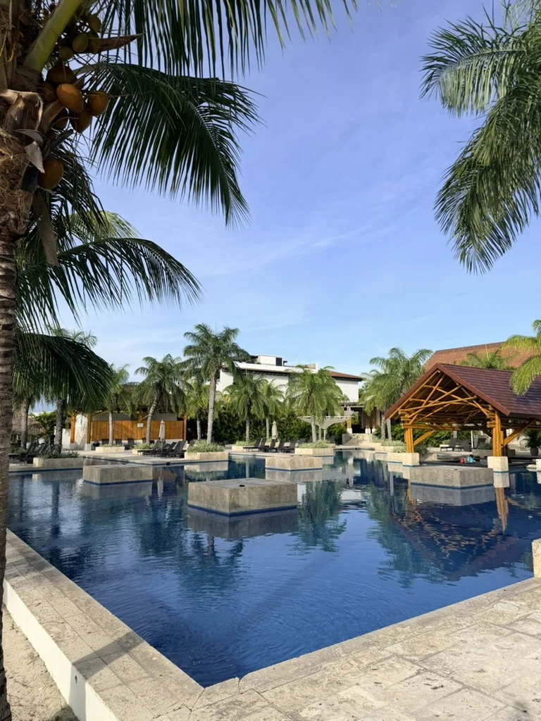 Large resort pool with geometric square islands and calm blue water. Palm trees and a gazebo structure in the background.